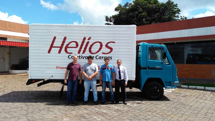 Foto de um veículo 3x4 com carroceria baú na cor branca e logo da Helios em vermelho, com cabina azul. À frente funcionários e associados da empresa da cidade de Santo Antonio do Sudoeste/PR.